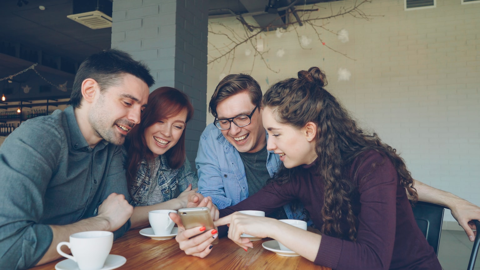 Four friends at a cafe table laughing while looking at a smartphone together